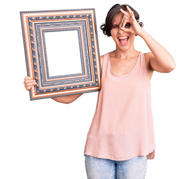Beautiful young woman with short hair holding empty frame smiling happy doing ok sign with hand on eye looking through fingers