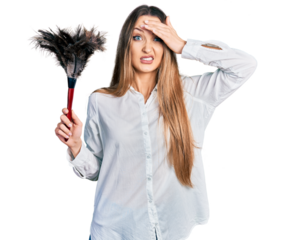 Beautiful caucasian woman holding cleaning duster stressed and frustrated with hand on head, surprised and angry face