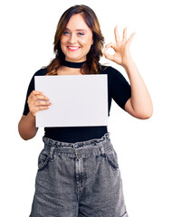 Young beautiful caucasian woman holding blank empty paper doing ok sign with fingers, smiling friendly gesturing excellent symbol