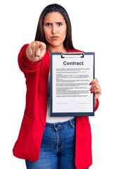Young beautiful brunette woman holding clipboard with contract pointing with finger to the camera and to you, confident gesture looking serious