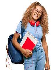 Beautiful caucasian teenager girl holding student backpack and books looking positive and happy...