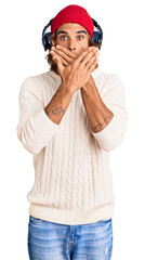 Young hispanic man listening to music using headphones shocked covering mouth with hands for mistake. secret concept.