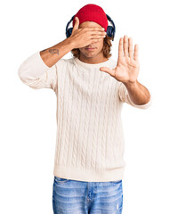 Young hispanic man listening to music using headphones covering eyes with hands and doing stop gesture with sad and fear expression. embarrassed and negative concept.