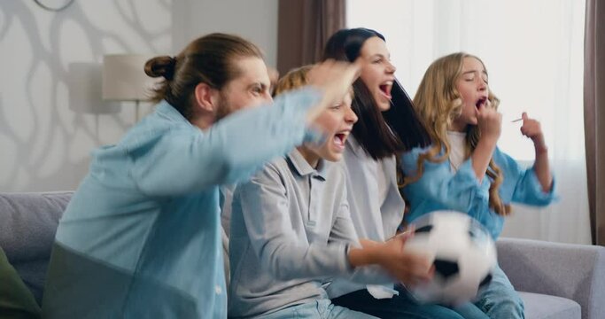 Family sitting on couch at home watching live broadcasting of soccer game together celebrating a goal. Happy parents, mom, dad and children with ball, supporting the victory of their favorite football