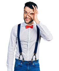 Hispanic man with beard wearing hipster look with bow tie and suspenders doing ok gesture with hand smiling, eye looking through fingers with happy face.