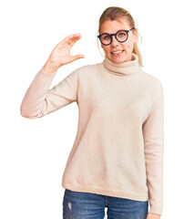 Young beautiful blonde woman wearing turtleneck sweater and glasses smiling and confident gesturing with hand doing small size sign with fingers looking and the camera. measure concept.