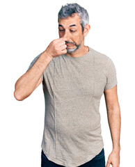 Middle age hispanic with grey hair wearing casual grey t shirt smelling something stinky and...