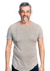 Middle age hispanic with grey hair wearing casual grey t shirt sticking tongue out happy with funny expression. emotion concept.