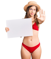 Beautiful caucasian young woman wearing bikini holding blank empty banner with open hand doing stop sign with serious and confident expression, defense gesture