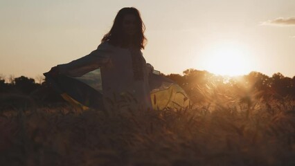 A Ukrainian girl in a dress walks holding a freely waving flag in a middle of the golden wheat crop field in the countryside. A young woman cherishing Ukraine's yellow and blue symbol in her homeland.