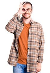 Young handsome caucasian man wearing casual clothes and glasses smiling happy doing ok sign with hand on eye looking through fingers