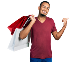 Young african american man holding shopping bags pointing thumb up to the side smiling happy with open mouth
