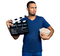 Young african american man eating popcorn holding film clapboard in shock face, looking skeptical...
