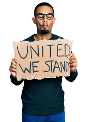 Young african american man holding united we stand banner making fish face with mouth and squinting eyes, crazy and comical.