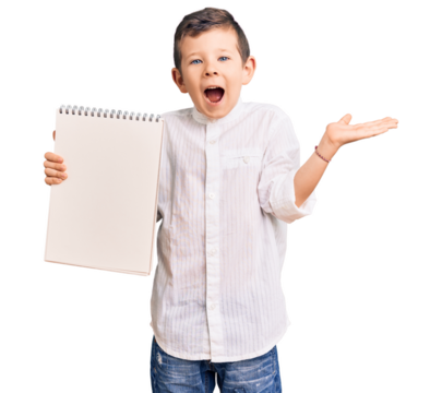 Cute blond kid holding notebook celebrating victory with happy smile and winner expression with raised hands