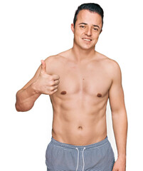 Fototapeta premium Handsome young man wearing swimwear shirtless doing happy thumbs up gesture with hand. approving expression looking at the camera showing success.