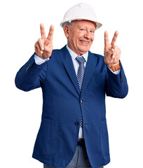 Senior handsome grey-haired man wearing suit and architect hardhat smiling looking to the camera showing fingers doing victory sign. number two.
