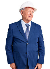 Senior handsome grey-haired man wearing suit and architect hardhat smiling looking to the side and staring away thinking.