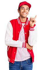 Young african amercian man wearing baseball uniform with a big smile on face, pointing with hand and finger to the side looking at the camera.
