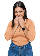 Young hispanic woman wearing casual clothes laughing and embarrassed giggle covering mouth with...