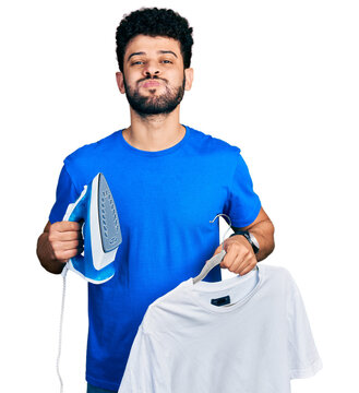 Young arab man with beard holding electric steam iron and white t shirt puffing cheeks with funny face. mouth inflated with air, catching air.