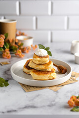A stack of lush, ruddy yellow-brown homemade pancakes with topping - white sauce and fresh mint greens in a white plate on the table with drinks and ingredients for a delicious meal.                

