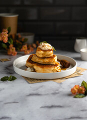 A stack of lush, ruddy yellow-brown homemade pancakes with topping - white sauce, cinnamon, honey and fresh mint greens in a white plate on the table with drinks and ingredients for a delicious meal.