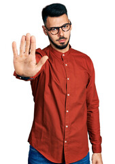 Young hispanic man with beard wearing business shirt and glasses doing stop sing with palm of the hand. warning expression with negative and serious gesture on the face.