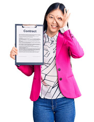 Young beautiful chinese woman holding clipboard with contract document smiling happy doing ok sign with hand on eye looking through fingers