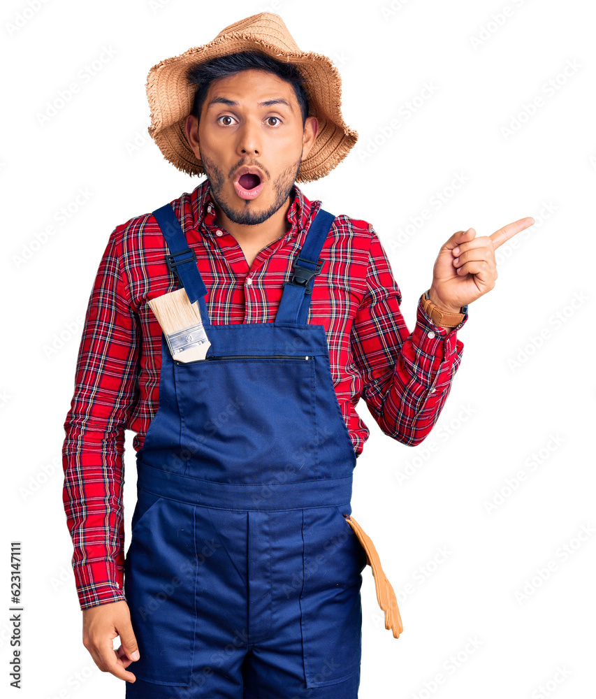 Wall mural Handsome latin american young man weaing handyman uniform surprised pointing with finger to the side, open mouth amazed expression.