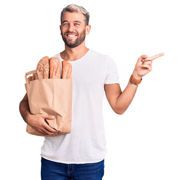 Young handsome blond man holding paper bag with bread smiling happy pointing with hand and finger to the side