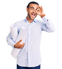 Young handsome man holding paper blueprints smiling happy doing ok sign with hand on eye looking through fingers