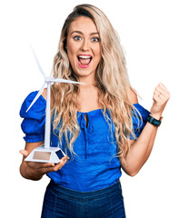 Young blonde woman holding solar windmill for renewable electricity pointing thumb up to the side smiling happy with open mouth