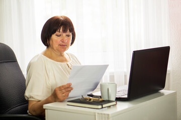 Middle aged senior woman sitting with laptop and paper document, working and drinking coffee.
