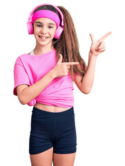 Obraz na płótnie Canvas Cute hispanic child girl wearing gym clothes and using headphones smiling and looking at the camera pointing with two hands and fingers to the side.