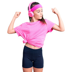 Fototapeta na wymiar Cute hispanic child girl wearing gym clothes and using headphones showing arms muscles smiling proud. fitness concept.