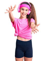 Obraz na płótnie Canvas Cute hispanic child girl wearing gym clothes and using headphones looking at the camera smiling with open arms for hug. cheerful expression embracing happiness.