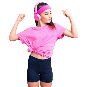 Cute hispanic child girl wearing gym clothes and using headphones showing arms muscles smiling proud. fitness concept.