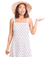 Young beautiful chinese girl wearing summer hat smiling cheerful presenting and pointing with palm of hand looking at the camera.