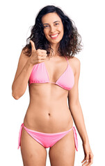 Young beautiful hispanic woman wearing bikini doing happy thumbs up gesture with hand. approving expression looking at the camera showing success.