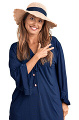 Middle age hispanic woman wearing summer hat cheerful with a smile of face pointing with hand and finger up to the side with happy and natural expression on face