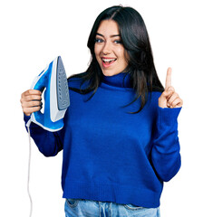 Beautiful hispanic woman holding electric steam iron smiling with an idea or question pointing finger with happy face, number one
