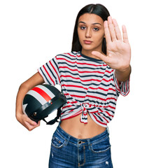 Young hispanic girl holding motorcycle helmet with open hand doing stop sign with serious and...