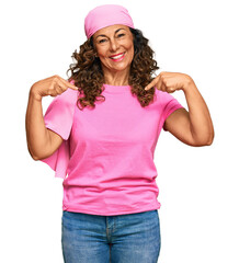 Middle age hispanic woman wearing breast cancer support pink scarf looking confident with smile on face, pointing oneself with fingers proud and happy.