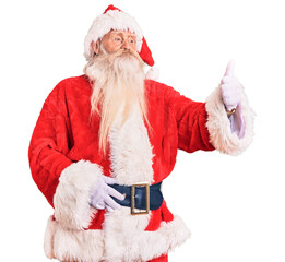 Old senior man with grey hair and long beard wearing traditional santa claus costume looking proud, smiling doing thumbs up gesture to the side