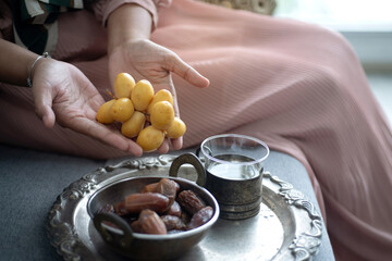 Hands of Muslim woman siting on couch and holding fresh dates fruit and dried dates in metal bowl nearby in tray, Iftar meal, Ramadan Kareem fasting month concept