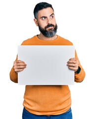 Hispanic man with beard holding blank empty banner smiling looking to the side and staring away thinking.