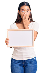 Young beautiful latin girl holding empty white chalkboard covering mouth with hand, shocked and afraid for mistake. surprised expression