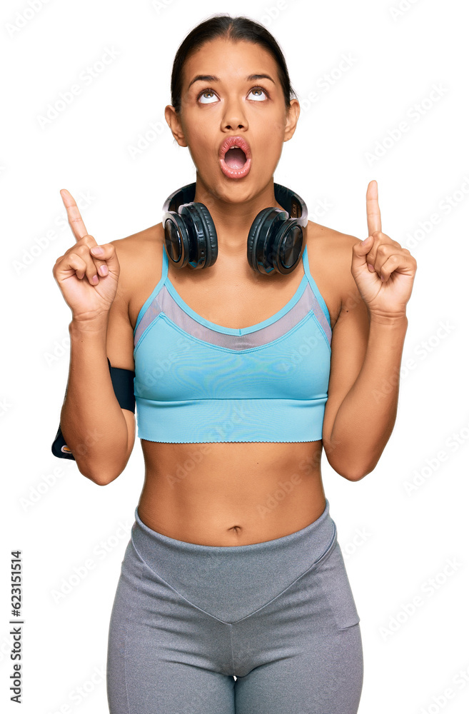 Sticker beautiful hispanic woman wearing sportswear and headphones amazed and surprised looking up and point