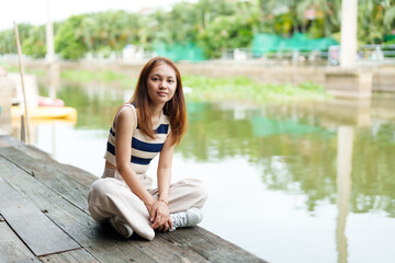Fototapeta na wymiar Cute Asian girl with long hair wearing short sleeves Sit and relax in spare time at the pier. By the river on weekends with a smiling face A rural canal with clear water